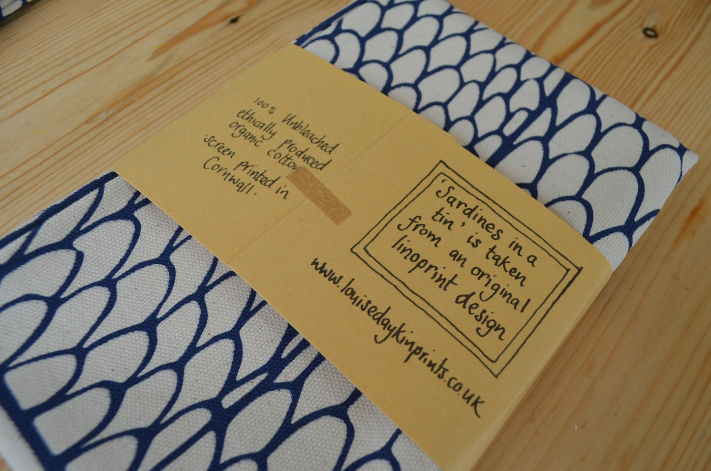 Image shows the back of the wrapped "Sardines in a Tin" Tea towel. The packaging reads "100% Unbleached ethically produced organic cotton. Screen printed in Cornwall. "Sardines in a tin" is taken from an original linoprint design. www.louisedaykinprints.co.uk
