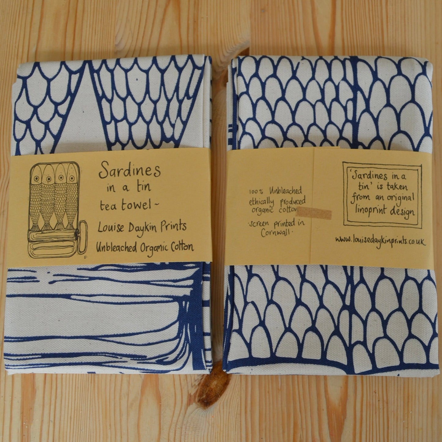 Image shows the front and back of the wrapped "Sardines in a Tin" Tea towel. Front handwritten text reads "Sardines in a tin tea towel- Louise Daykin Prints, Unbleached Organic Cotton. Back of the package reads "100% Unbleached ethically produced organic cotton. Screen printed in Cornwall. "Sardines in a tin" is taken from an original linoprint design. www.louisedaykinprints.co.uk
