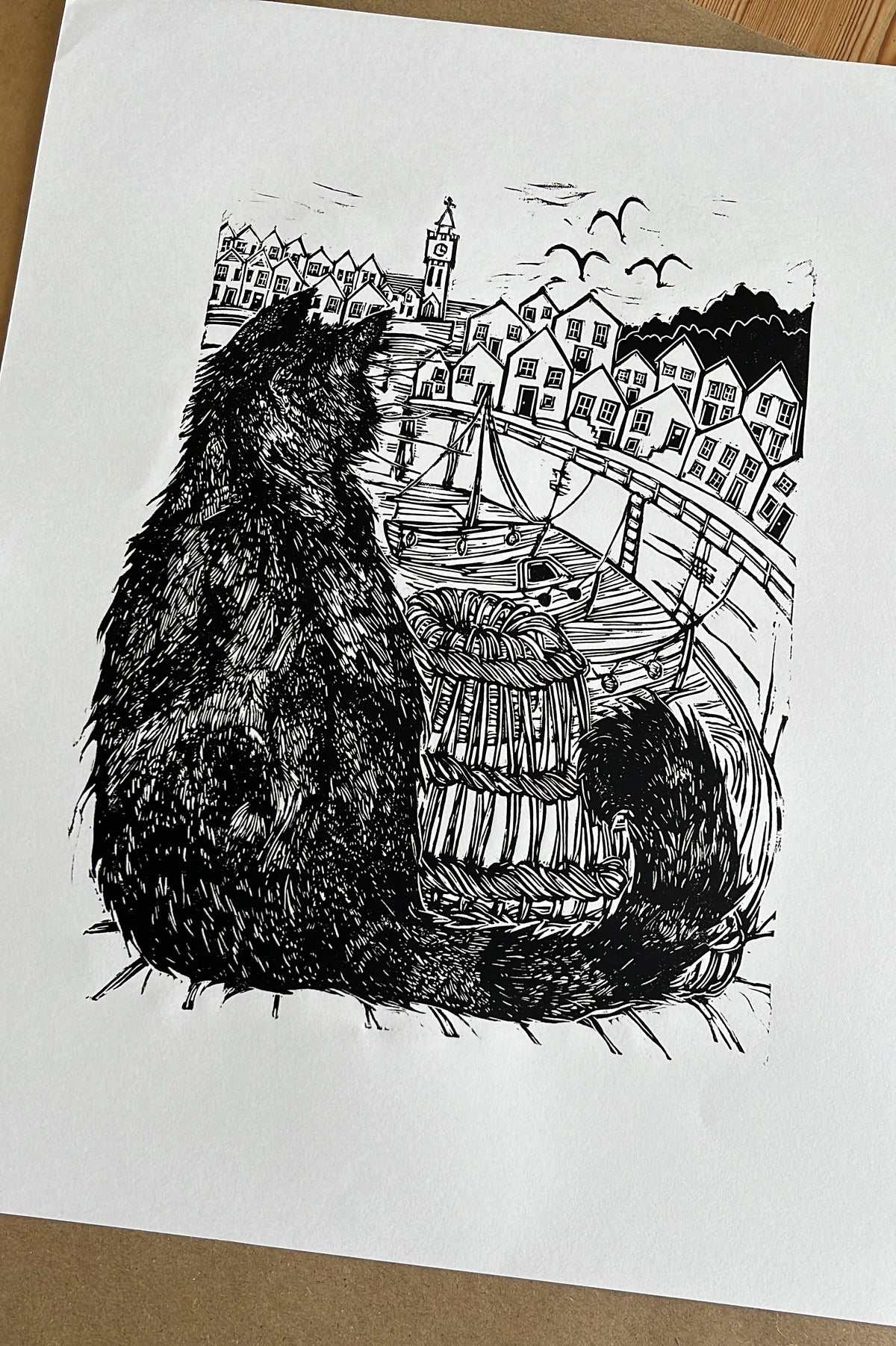 A black and white original limited edition linocut art print depicting a cat looking out over a Cornish harbour town, displayed on a brown kraft paper background.