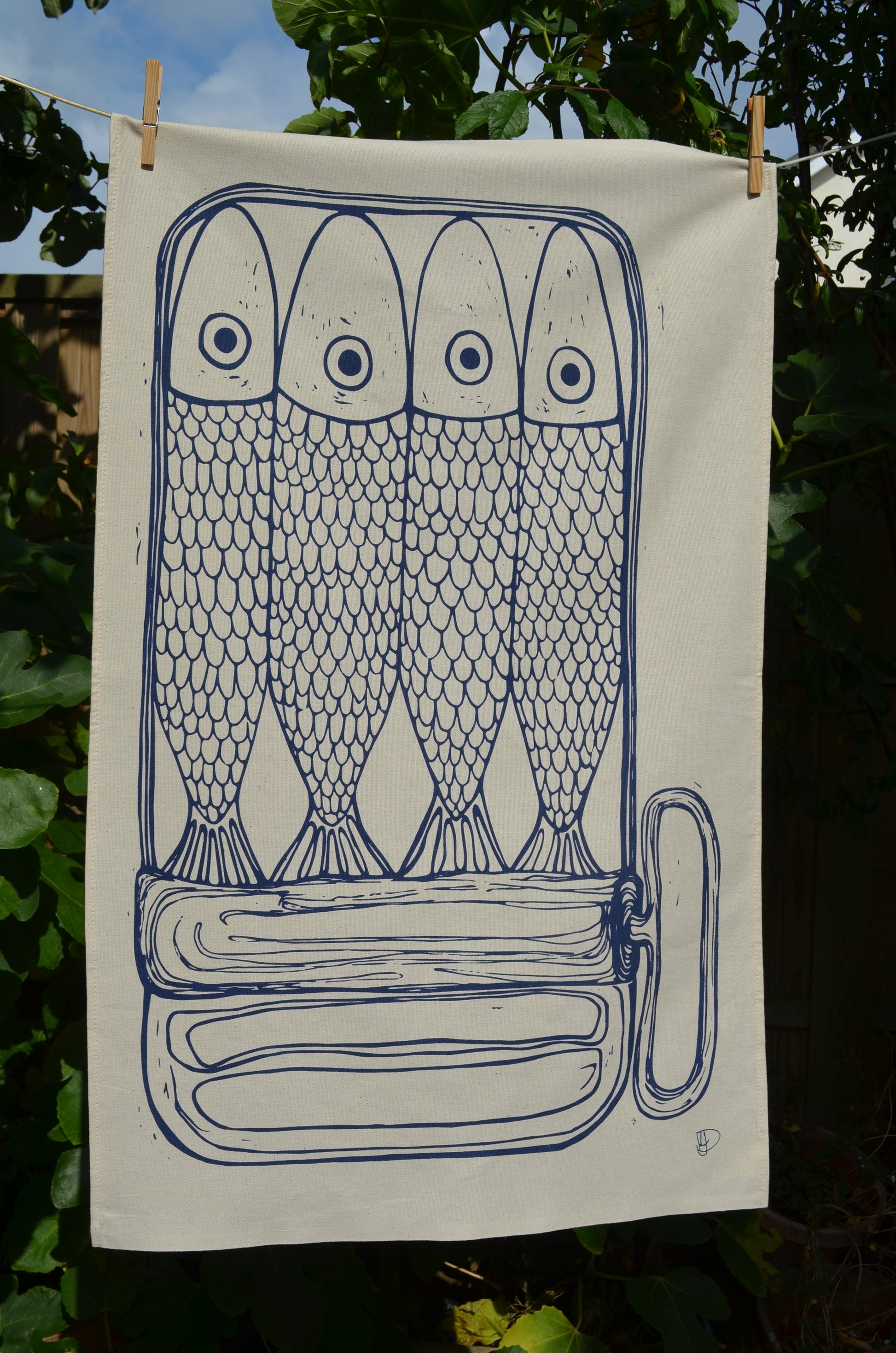 "Sardines in a Tin" full tea towel hanging in the sunshine. The design shows 4 sardines and their scales with the tin rolled down to a three quarter view so you can see the upper part of their tails.